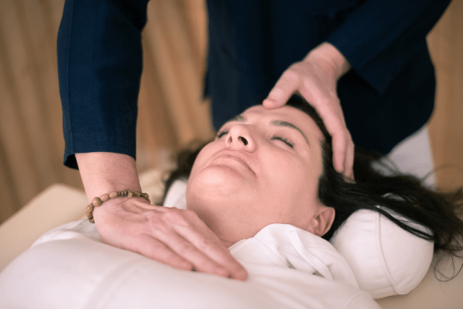 Craniosacral therapy practitioner resting one hand gently on a client's forehead and the other at the base of the neck while the client lies relaxed on a treatment table